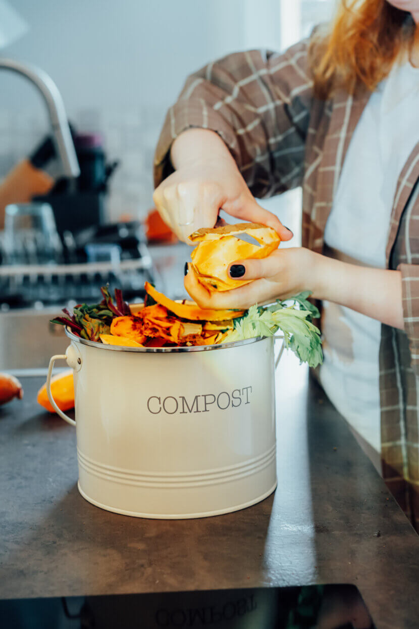 Kompostieren in der Wohnung: Funktioniert das wirklich? Compost the kitchen waste, recycling at home. Compost bin with vegetables cutted leftovers on the kitchen table with blurred woman cooking meal. Environmentally responsible, ecology. Selective focus